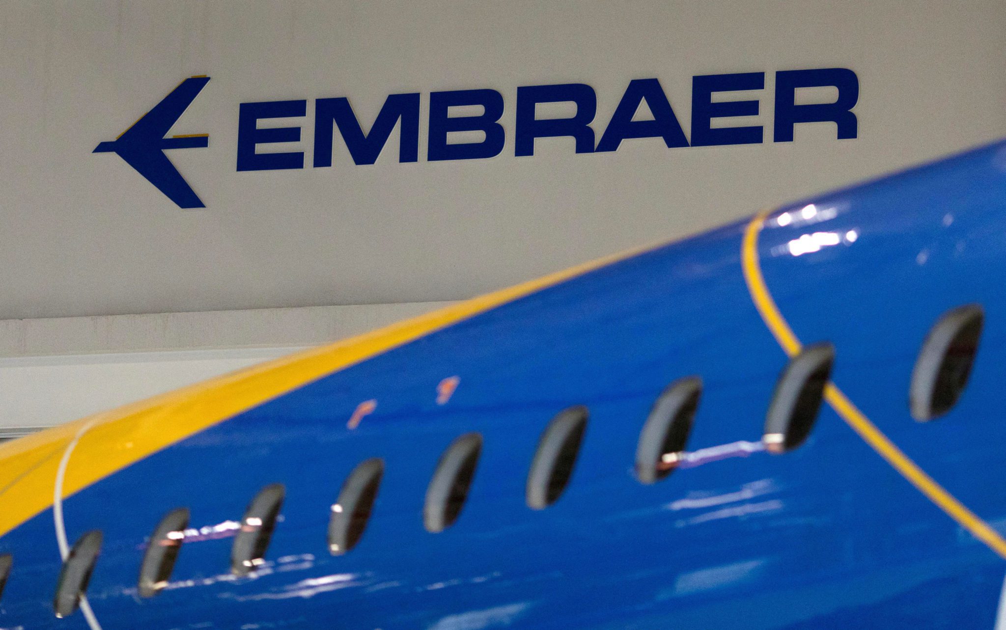 Logo azul da Embraer em parede branca, com parte da fuselagem de um avião azul e amarelo em primeiro plano.