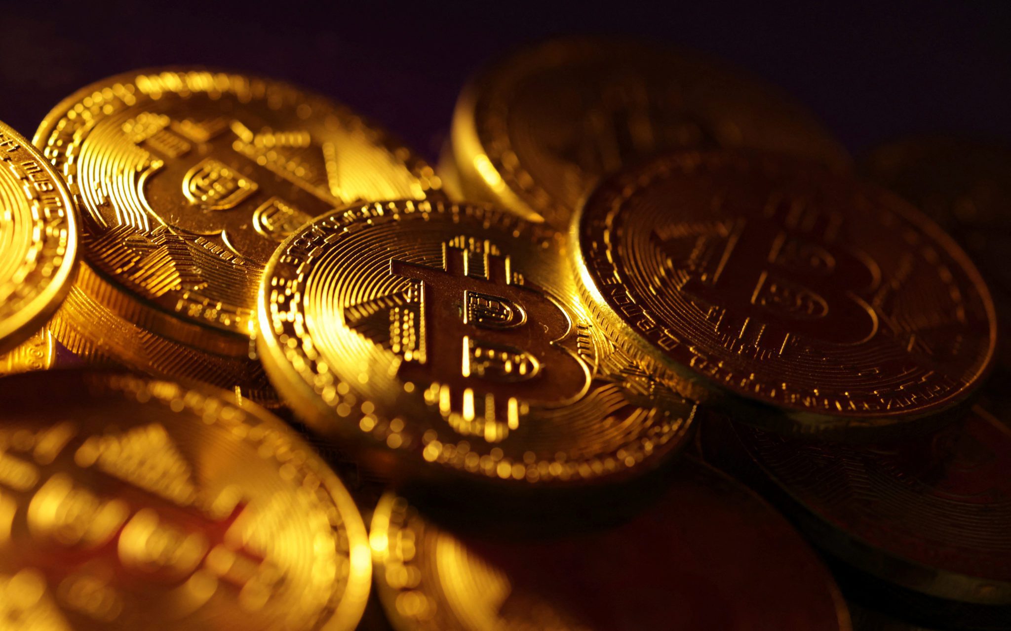 Várias moedas douradas de Bitcoin empilhadas, com o símbolo 'B' brilhando sob luz intensa.