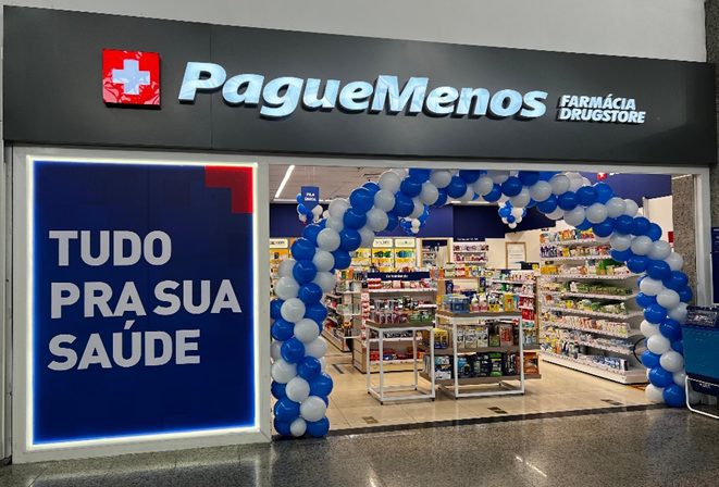 Entrada da Pague Menos Farmácia Drugstore com balões azul/brancos e cartaz 'Tudo pra sua saúde'.