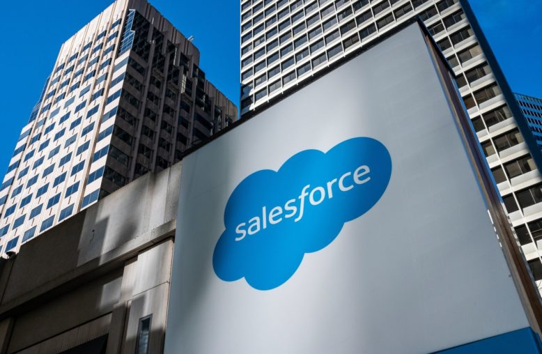 Sinal grande branco com o logo azul da Salesforce e edifícios modernos ao fundo.