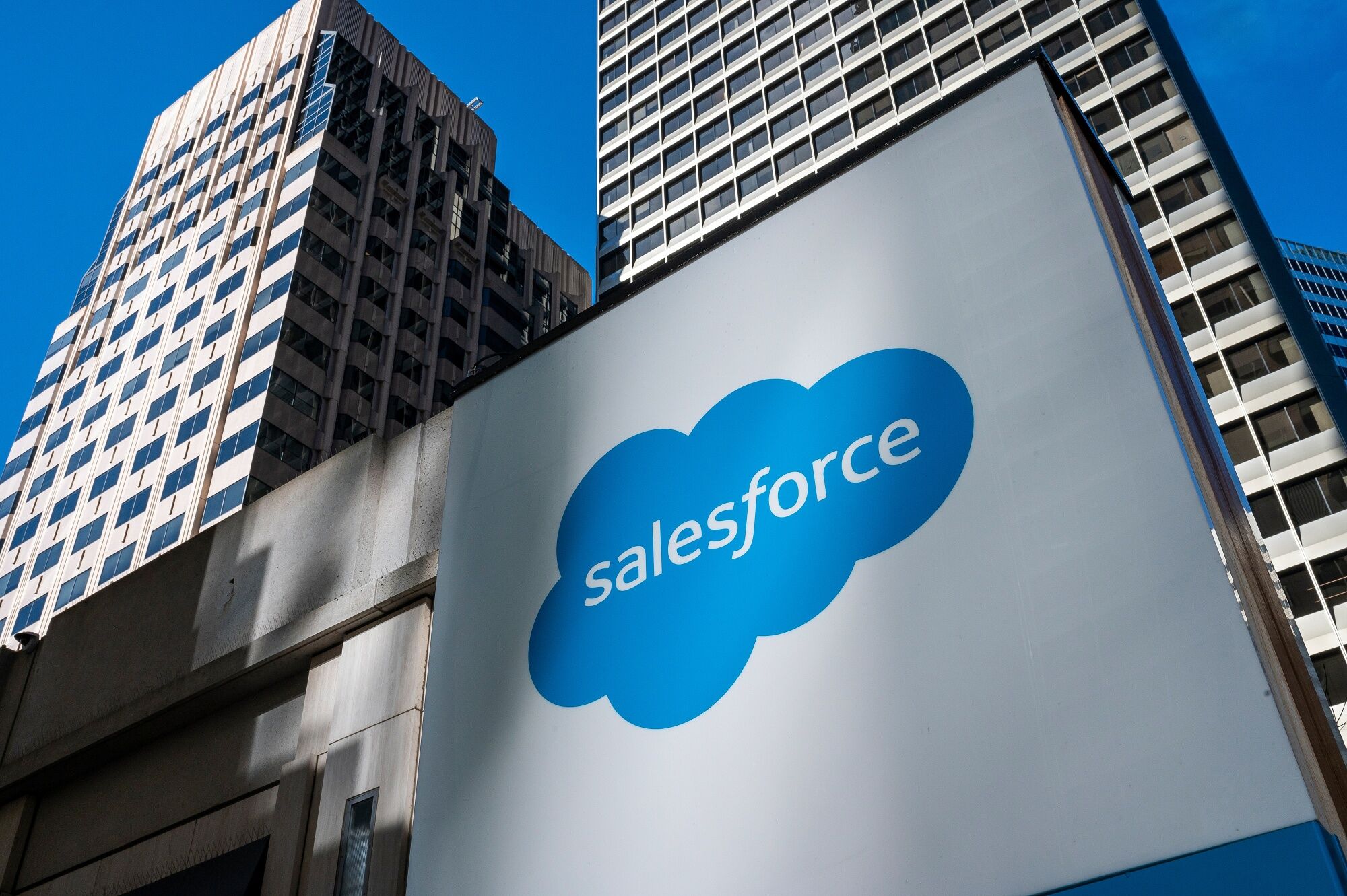 Sinal grande branco com o logo azul da Salesforce e edifícios modernos ao fundo.