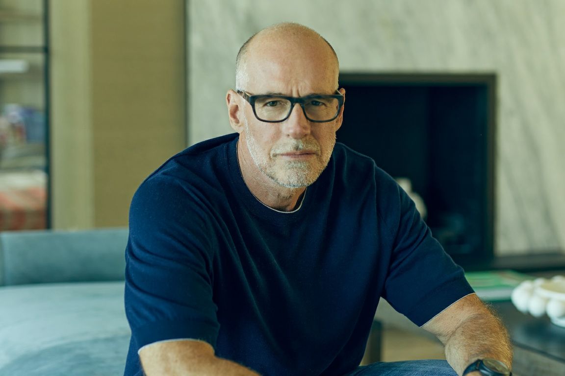 Homem careca de barba grisalha e óculos, com camisa azul, sentado, olhando fixamente para a câmera.