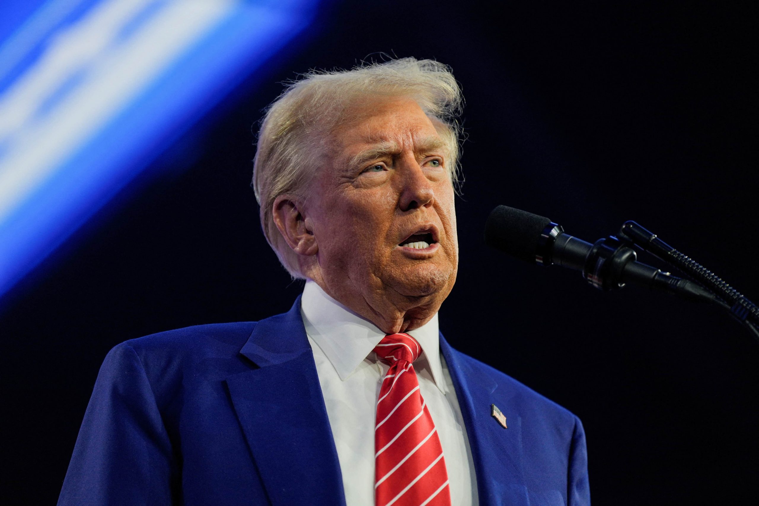 Donald Trump, em close-up, de terno azul e gravata vermelha, discursa ao microfone.