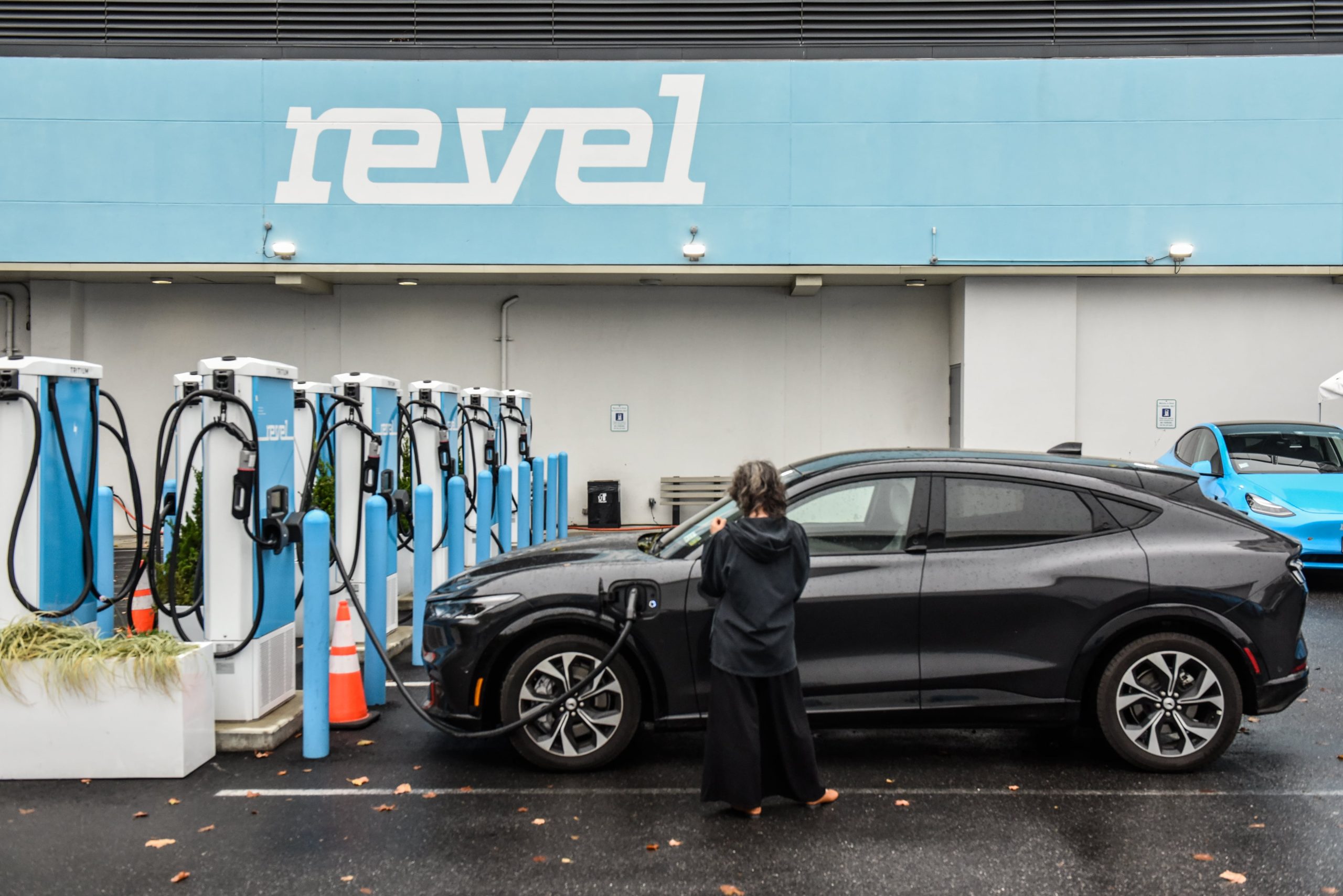 Pessoa carregando carro elétrico preto em estação Revel. Vários carregadores e um carro azul ao fundo.