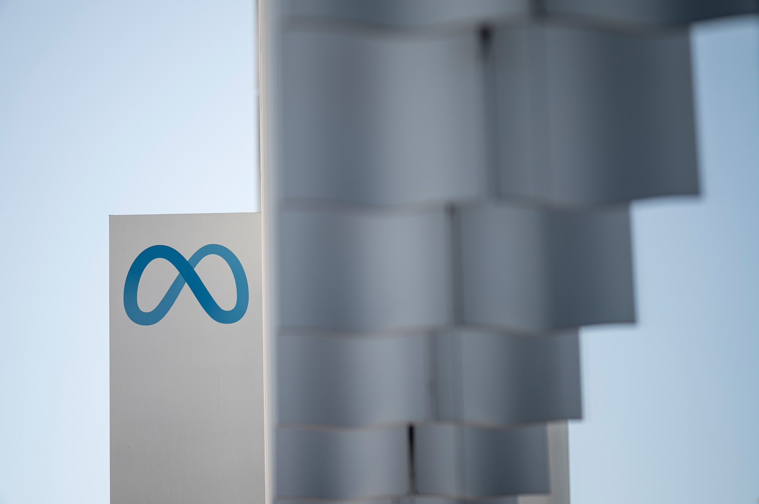 Logotipo azul do infinito da Meta em letreiro branco, com céu claro e estrutura desfocada ao lado.