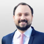 Retrato de um homem sorrindo com barba, camisa branca, terno azul e gravata rosa.