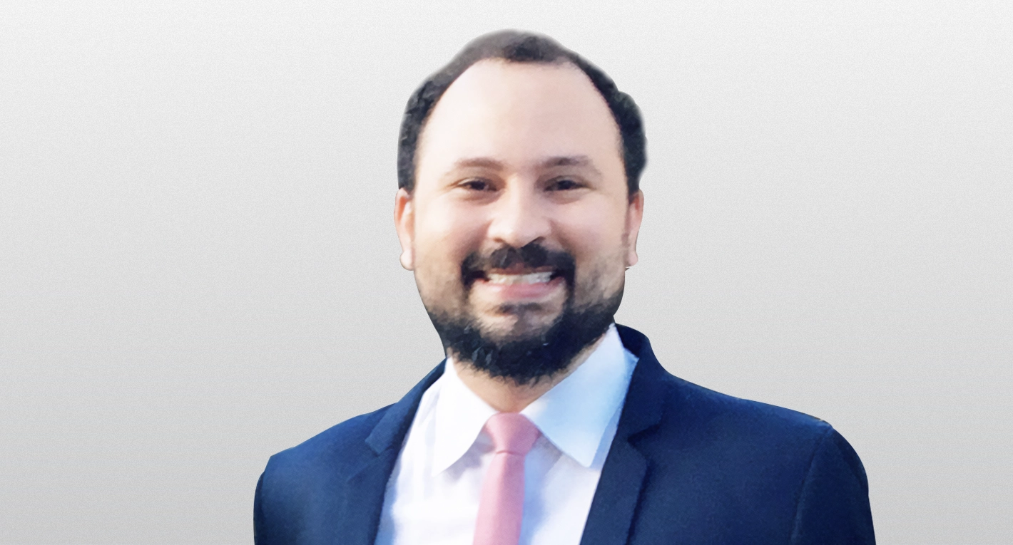 Retrato de um homem sorrindo com barba, camisa branca, terno azul e gravata rosa.