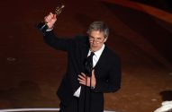 Homem de terno e óculos ergue um Oscar com a mão direita e coloca a esquerda no peito, no palco.