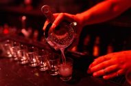 Barman coa e derrama bebida em shot, com copos na barra sob luz vermelha.