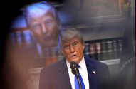 Donald Trump fala ao microfone. Ao fundo, uma projeção borrada de seu rosto e estantes de livros.