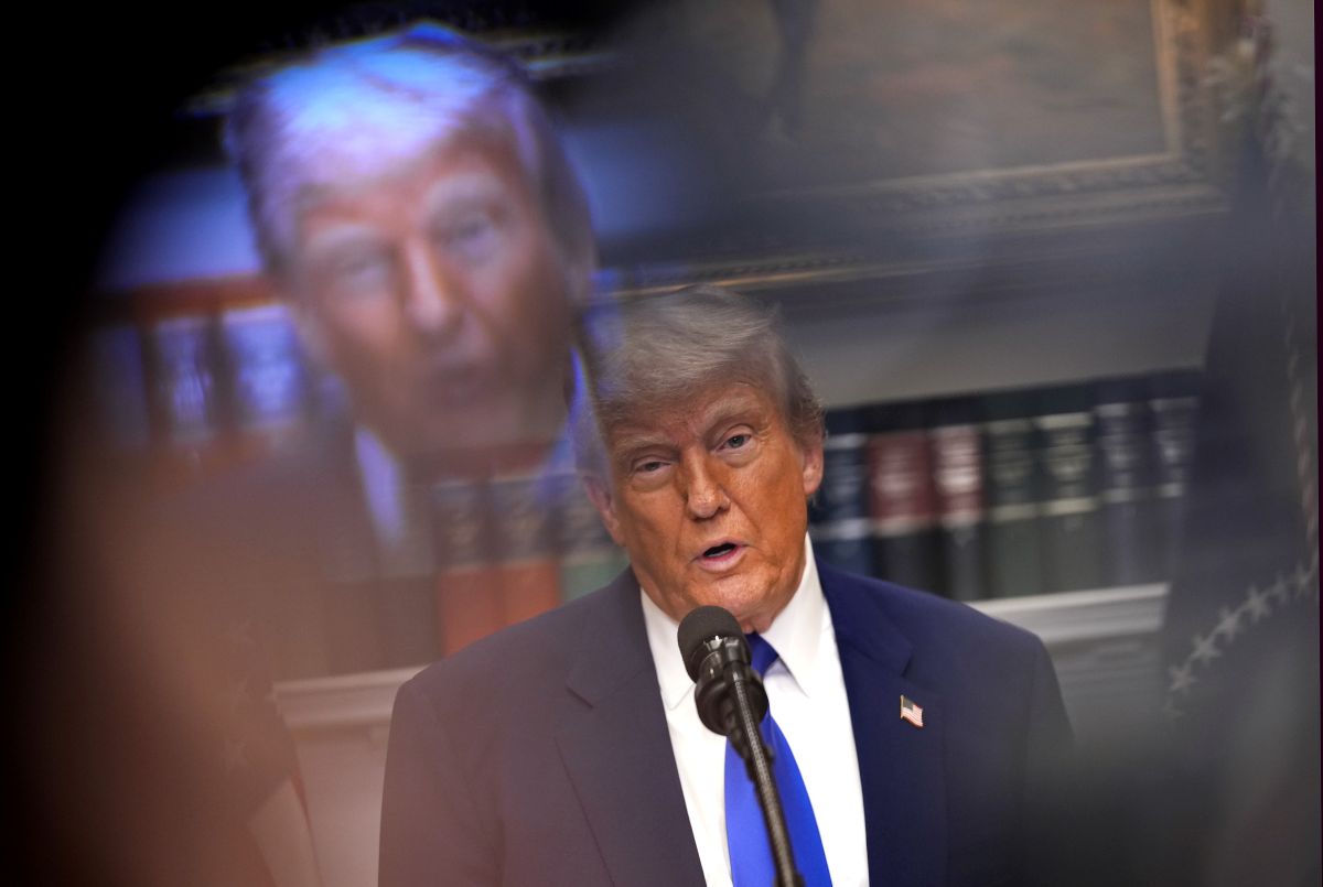 Donald Trump fala ao microfone. Ao fundo, uma projeção borrada de seu rosto e estantes de livros.