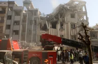 Edifício parcialmente desabado, fumaça. Caminhão de bombeiros com escada estendida. Equipes de resgate e multidão.