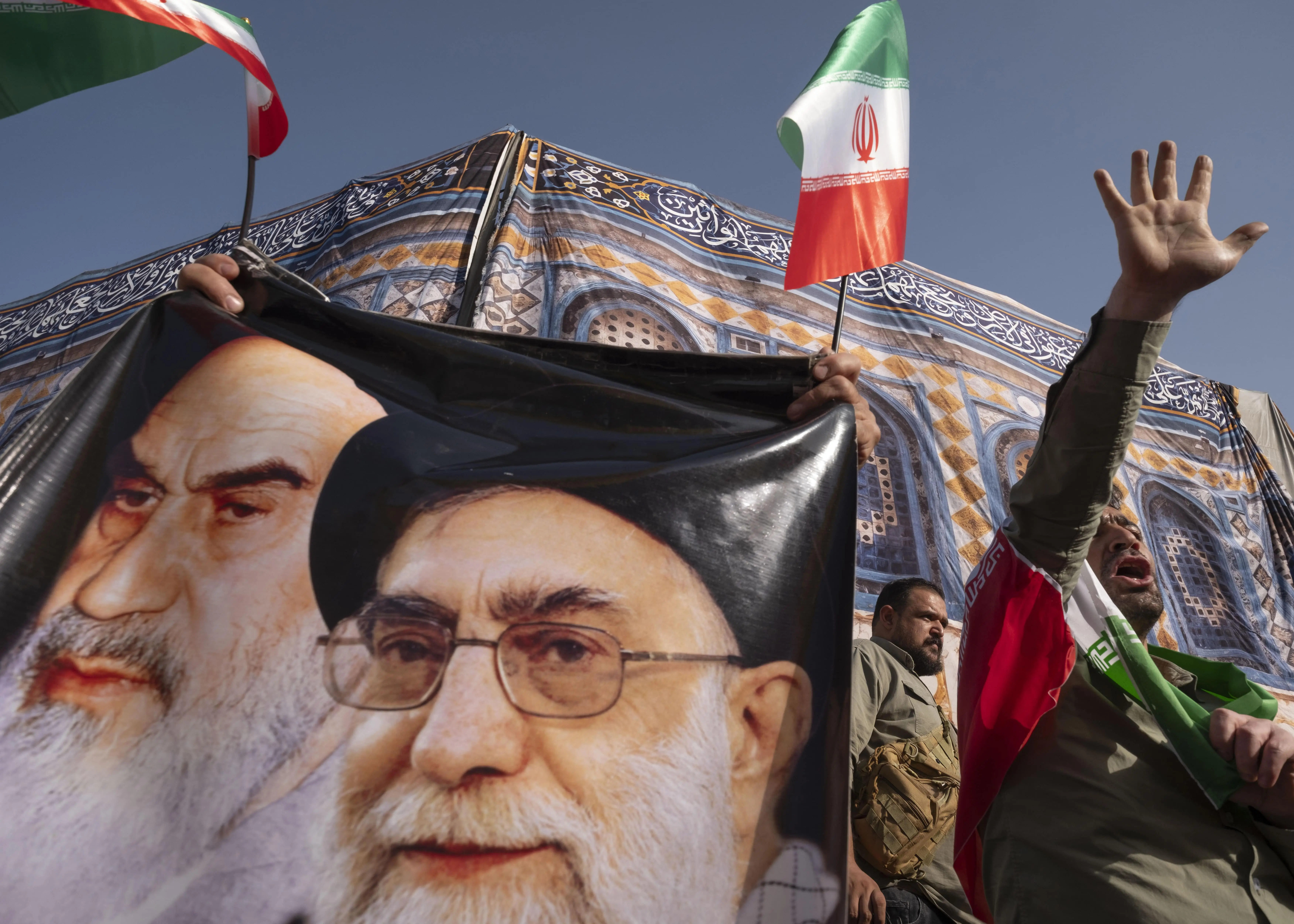 Pessoas seguram banner de Khomeini e Khamenei com bandeiras do Irã; um participante levanta o braço.