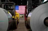 Duas grandes bobinas de metal em fábrica, ladeando empilhadeiras e uma bandeira dos EUA ao fundo.