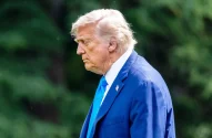 Donald Trump em terno azul e gravata, em perfil, olhando para baixo. Fundo borrado verde.