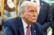 Donald Trump, de terno azul com pin da bandeira, sentado em um escritório. Ele tem um olhar sério e pensativo.