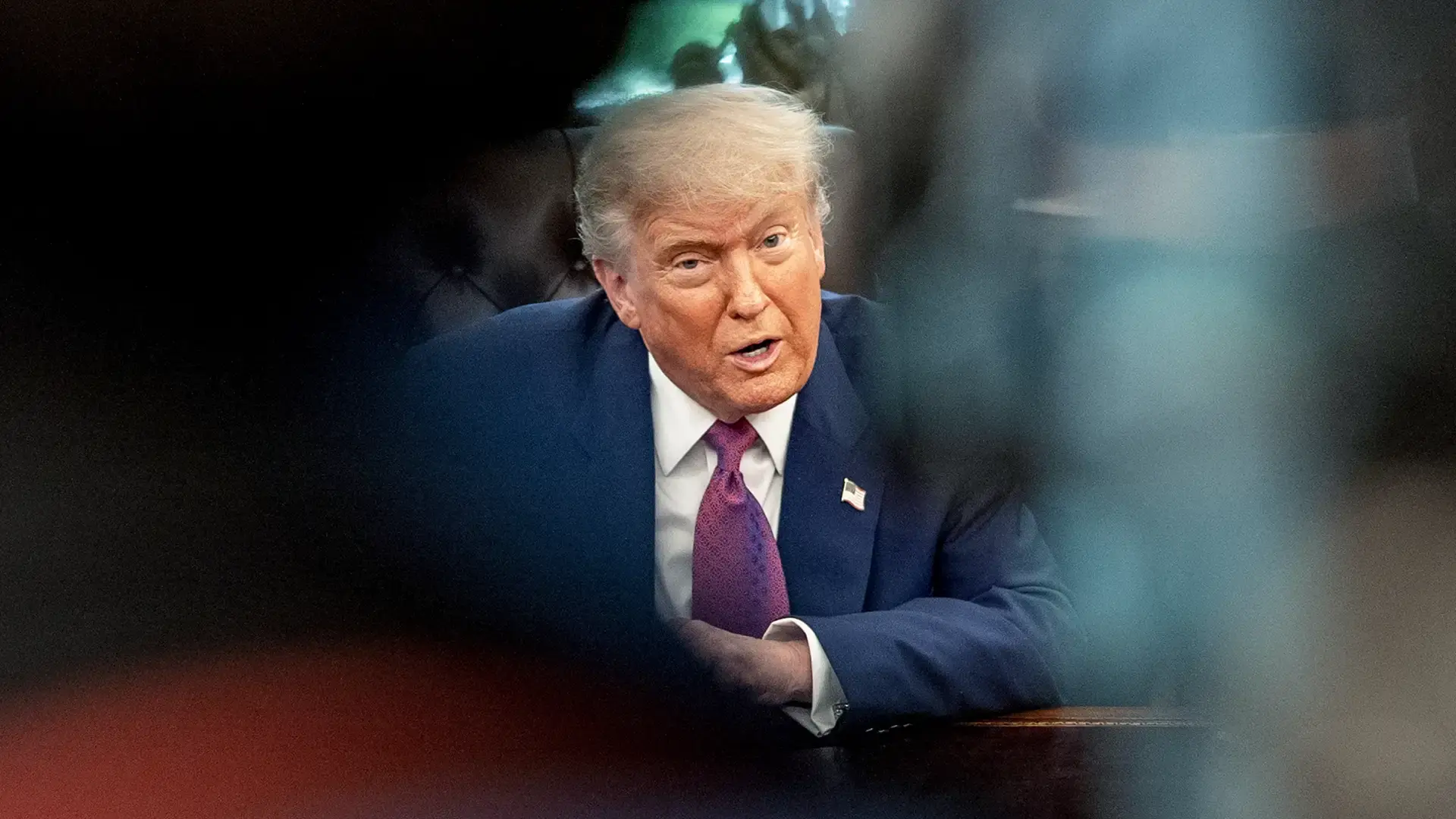 Donald Trump de terno, sentado em um escritório, falando. A imagem é enquadrada por elementos escuros e desfocados.