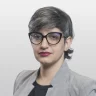 Mulher de cabelo grisalho curto, óculos e blazer xadrez cinza, olhando para a câmera.