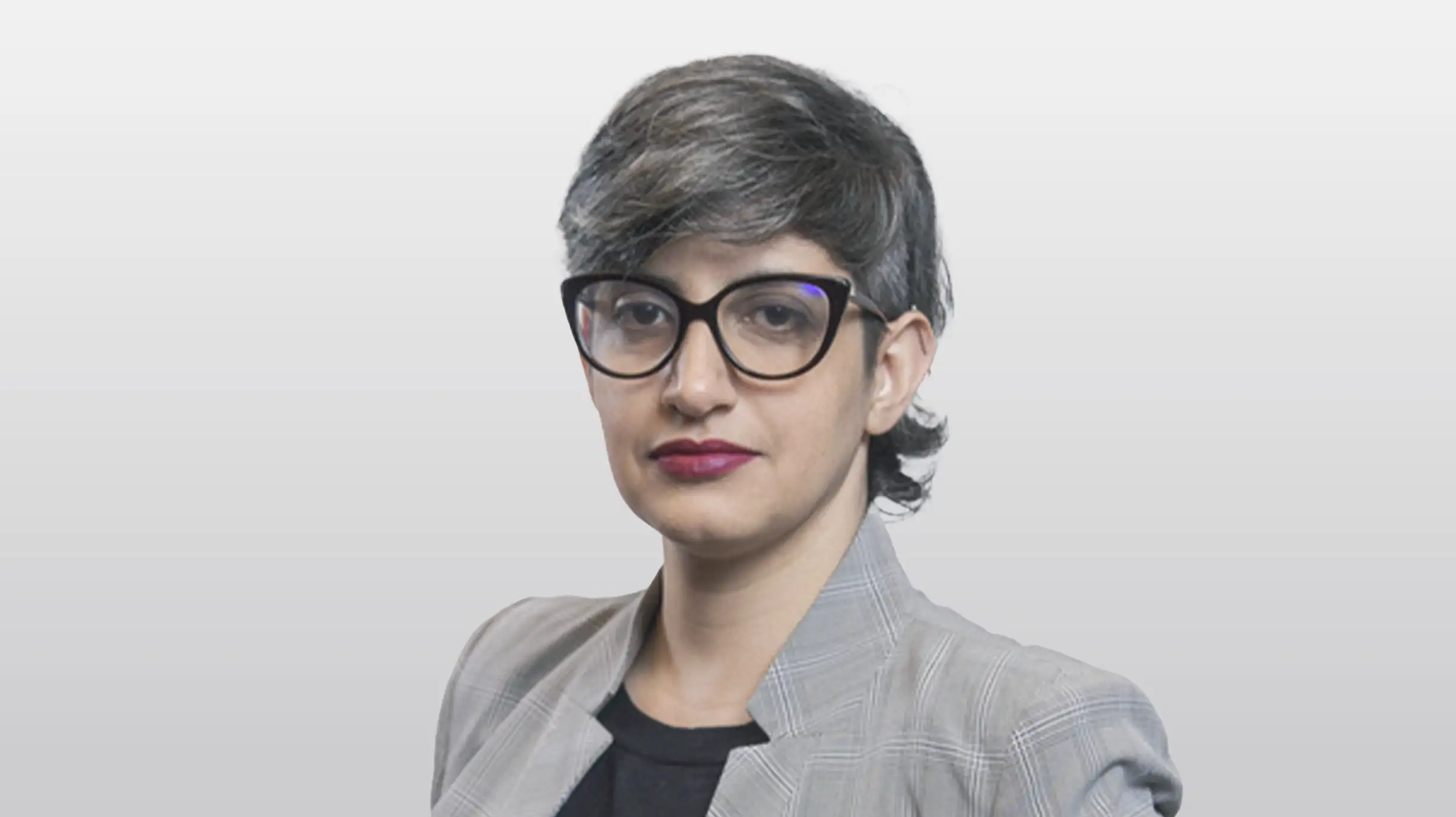 Mulher de cabelo grisalho curto, óculos e blazer xadrez cinza, olhando para a câmera.