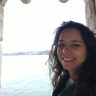 Mulher sorrindo em janela de pedra, com água e cidade no fundo.