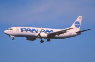 Avião Boeing 737 da Pan Am branco e azul em voo com trem de pouso abaixado, contra céu azul.